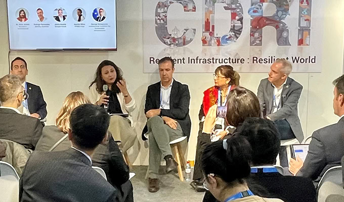 Panel discussion in progress with five panelists seated onstage, discussing infrastructure and resilience. Audience members are visible in the foreground, and a presentation slide is displayed.