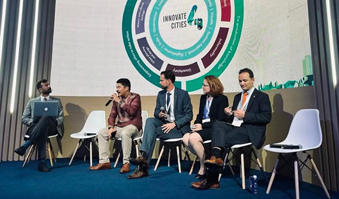 Five people are seated on a stage in front of a screen displaying a diagram labeled "Innovate Cities." One person is speaking passionately into a microphone about sustainable actions, while the others are attentively listening.