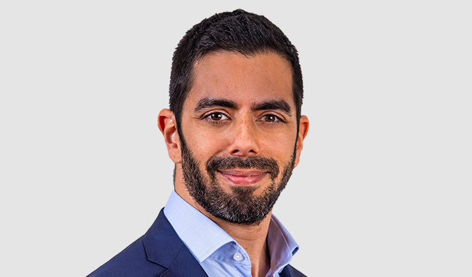 Un hombre con barba y cabello corto, vestido con un traje azul y una camisa azul claro, sonríe a la cámara sobre un fondo liso, encarnando el espíritu de la sostenibilidad.