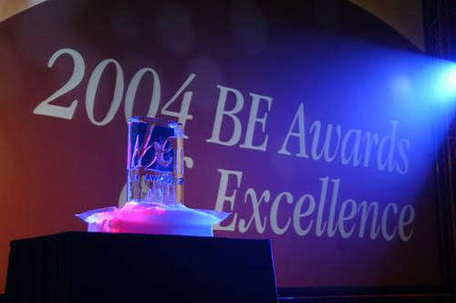 Statuetka na czarnym postumencie z napisem „2004 BE Awards of Excellence” wyświetlonym na czerwonym ekranie w tle.