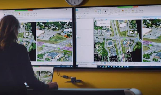 A person analyzes aerial maps displayed on two large monitors in a control room.