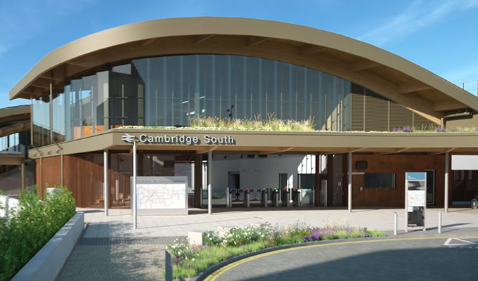 Modern building with arched roof and large glass windows, marked "Cambridge South," showcases sustainability in its design. The entrance features action-oriented ticket barriers and signage, surrounded by landscaped areas under a clear blue sky.