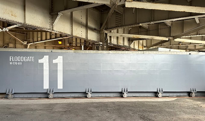 A large gray floodgate numbered 11 is positioned beneath an industrial steel structure, demonstrating a commitment to sustainability through action.