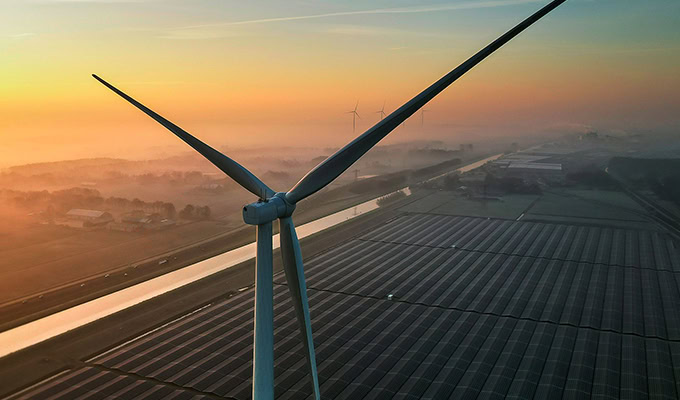 A wind turbine stands tall over a field of solar panels at sunrise, with a misty landscape and additional turbines visible in the distance.