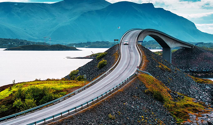 A curving bridge over water with a car on it, surrounded by green hills and mountains in the background.