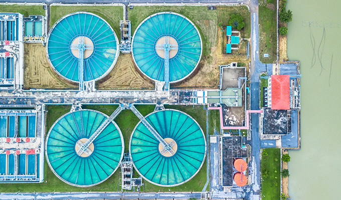 Aerial view of a wastewater treatment plant with four large circular tanks and several rectangular structures surrounded by pathways and greenery.