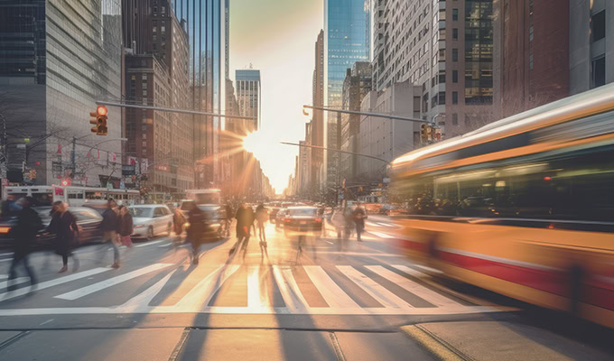 Intersezione di una strada cittadina trafficata al tramonto con edifici, pedoni che attraversano e movimento sfocato dei veicoli che passano.