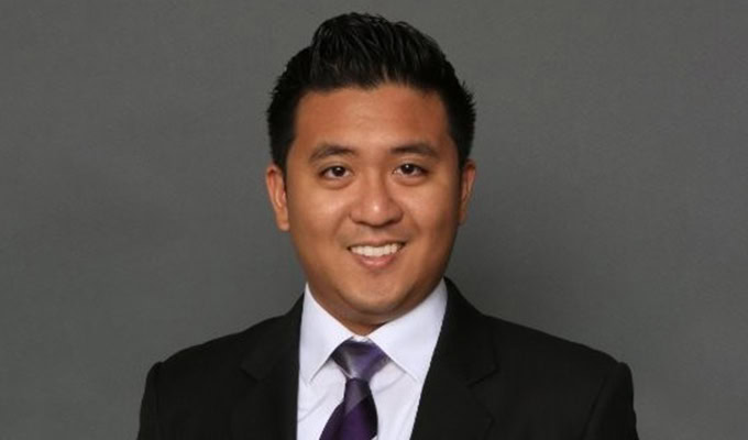 Un hombre con cabello corto y oscuro viste un traje negro, camisa blanca y corbata morada, sonriendo frente a un fondo gris liso.