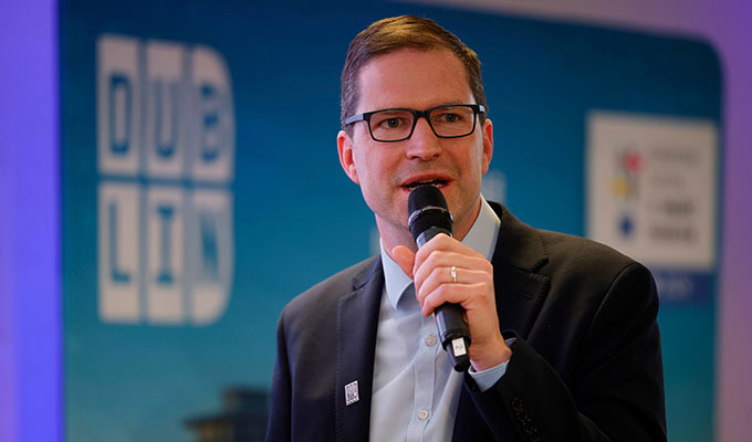 Ein Mann in schwarzem Anzug und Brille spricht in ein Mikrofon. Er steht vor einem Schild mit der Aufschrift "Dublin".