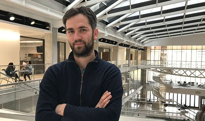 Um homem com barba e cabelos escuros está de braços cruzados em um átrio com grandes janelas e grades de vidro. As pessoas estão sentadas em mesas ao fundo.