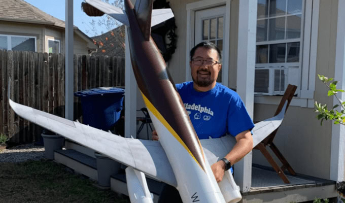 A man stands outside holding a large model airplane next to a small house with a wooden deck.