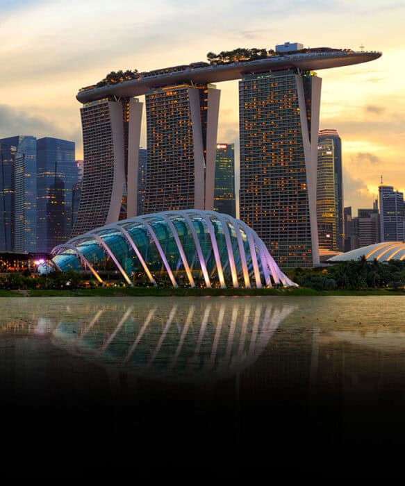 A stunning view of the Marina Bay Sands complex and the Flower Dome in Singapore during sunset, with their reflections visible in the water below. The Marina Bay Sands features three skyscrapers topped by a boat-shaped skypark, while the Flower Dome's illuminated glass structure curves gracefully in the foreground.