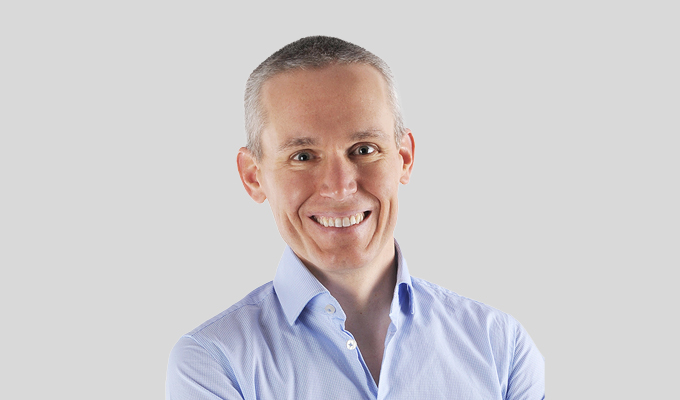 Person with short hair, smiling, wearing a light blue button-up shirt, posed against a plain gray background.