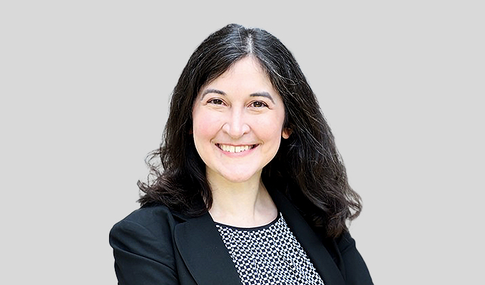 Smiling person with long dark hair wearing a black blazer over a patterned top, against a plain background.