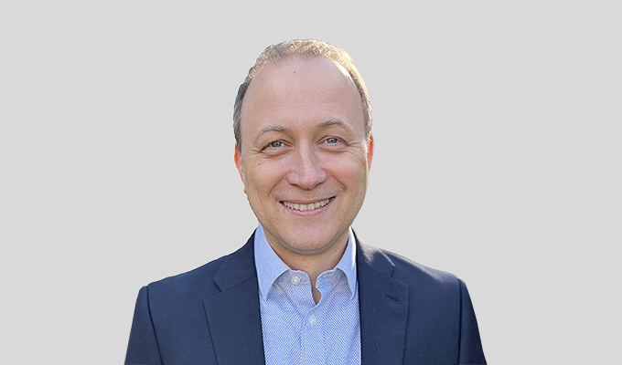 A smiling man in a suit and light blue shirt stands against a plain grey background.