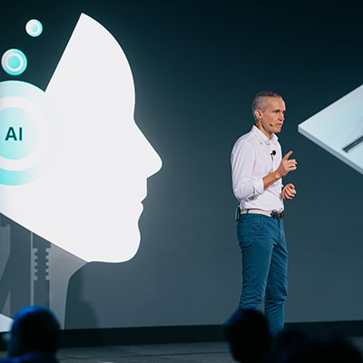 Um homem de camisa branca e calça azul fazendo uma apresentação no palco. Atrás dele, é exibido um gráfico grande de um perfil humano com “AI” escrito.