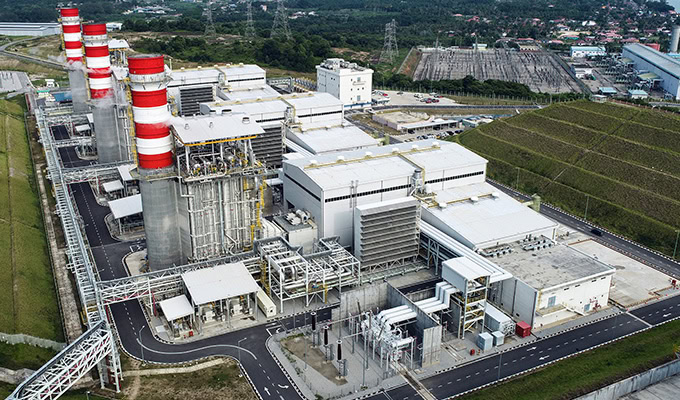 Vue aérienne d'une centrale électrique industrielle composée de plusieurs bâtiments, de cheminées et d'infrastructures environnantes, située à proximité d'une zone suburbaine et de vastes champs ouverts.