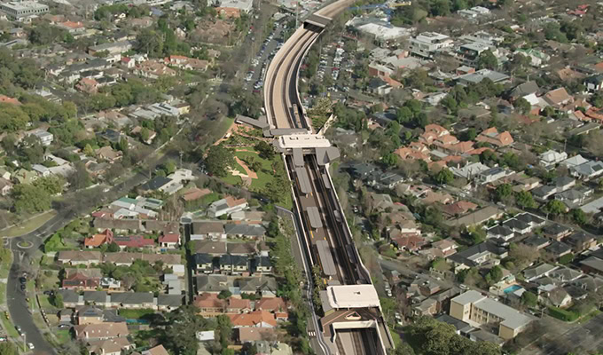 Vue aérienne d'une ligne ferroviaire surélevée traversant une zone résidentielle avec des maisons et des arbres de part et d'autre. Plusieurs bâtiments et routes sont visibles autour.