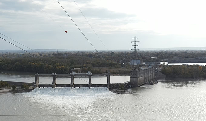 Duża zapora z wodą przepływającą przez jedną sekcję, otoczona wodami rzeki i krajobrazem z drzewami. Na pierwszym planie widoczne są linie energetyczne i czerwona kula.