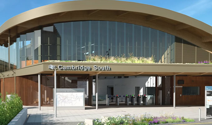 La imagen muestra la entrada de la estación de tren de Cambridge South con una arquitectura moderna de vidrio y madera, un techo arqueado y barreras de boletos visibles.