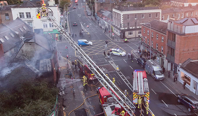 Einsatzkräfte der Feuerwehr löschen mit einem Leiterwagen einen Gebäudebrand in einer stark befahrenen Straße in der Stadt. Polizeiautos blockieren die Straße und Fußgänger schauen vom Bürgersteig aus zu.