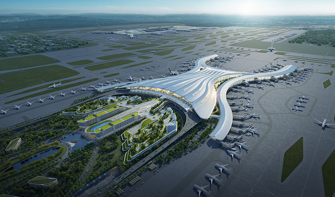 Aerial view of a large, modern airport terminal with multiple runways, taxiways, and airplanes positioned around the structure. Surrounding areas feature roads, parking facilities, and green spaces.