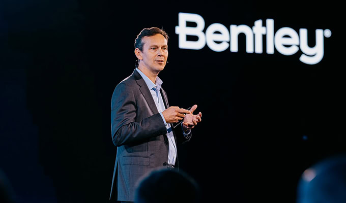 Person in a suit speaking on stage with a Bentley logo in the background.