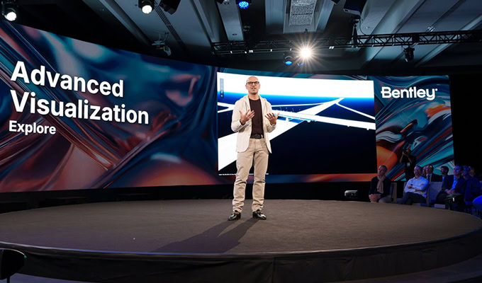 A person stands on stage presenting at an event with large screens displaying "Advanced Visualization Explore" and "Bentley.