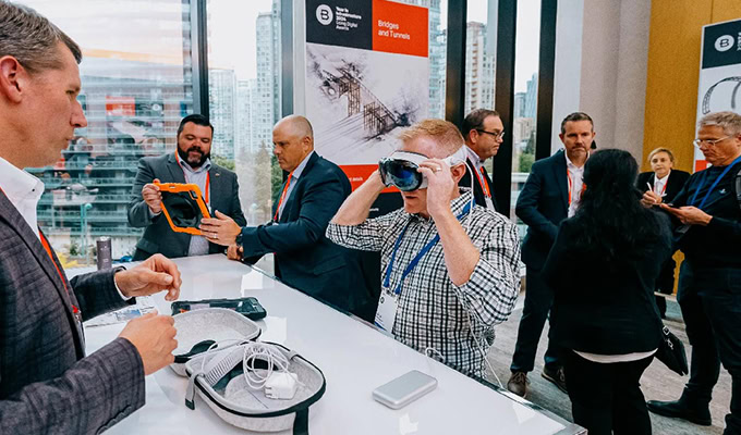 People at a tech conference, one individual trying on VR goggles, others interacting with gadgets at a booth. Large windows in the background display a cityscape.