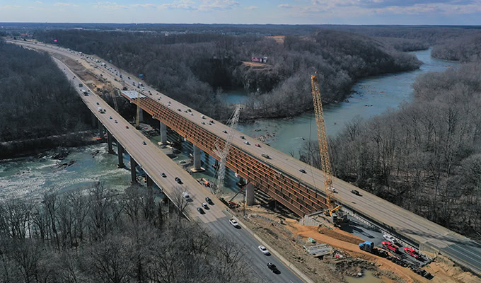 Luftaufnahme einer Baustelle an einem Fluss mit Kränen und Geräten, die an einer neuen Brücke parallel zu einer bestehenden Autobahnbrücke für den Verkehr arbeiten.