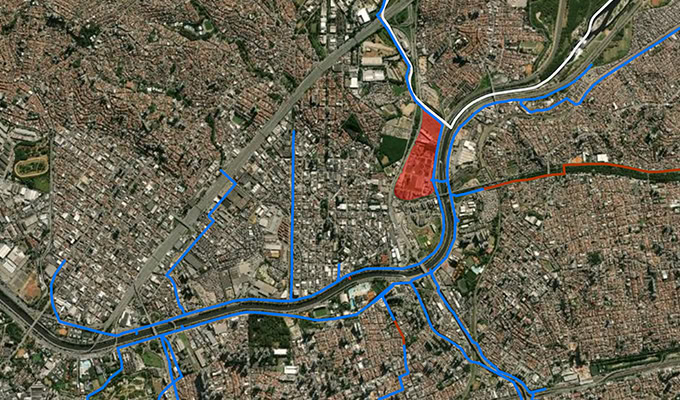 Aerial view of an urban area with various buildings and infrastructure. A river runs through the center, and blue and red lines mark specific boundaries and areas of interest.