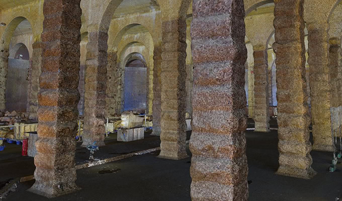 Un espace souterrain faiblement éclairé avec de multiples colonnes de pierre épaisses et nervurées et divers objets éparpillés sur le sol.