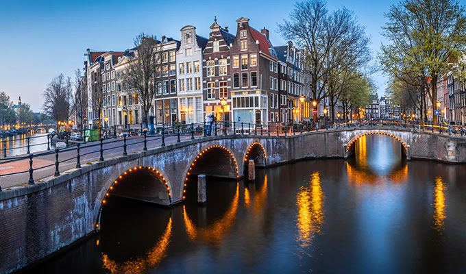 Un canale di Amsterdam al tramonto, con ponti ad arco illuminati da luci. Edifici storici fiancheggiano il corso d'acqua, riflettendo i tenui colori della sera.