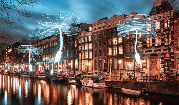 Leuchtende schirmartige Installationen schweben in der Abenddämmerung über einer von Gebäuden und Booten gesäumten Gracht und spiegeln sich im Wasser.