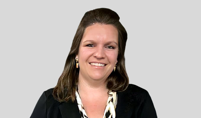 A woman with shoulder-length brown hair, wearing a black blazer over a patterned top, smiles against a plain, light gray background.