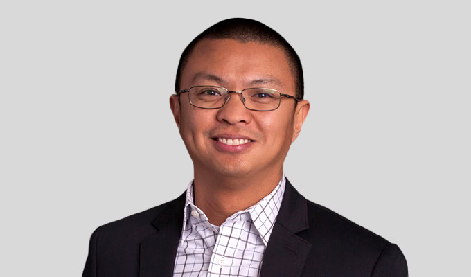 A person wearing glasses, a dark blazer, and a checkered shirt smiling against a plain background.