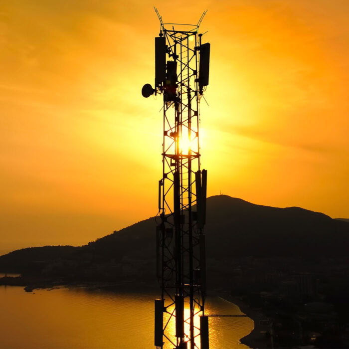 Uma torre de telecomunicações aparece em silhueta contra um pôr do sol alaranjado, com água e colinas ao fundo, incorporando a essência do OpenTower®.