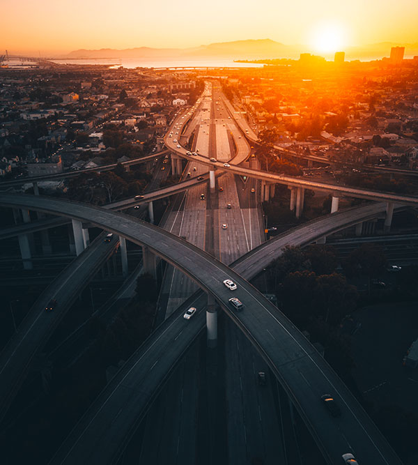 An aerial view of a sprawling highway interchange highlights the intricate transportation network against a sunset backdrop over a cityscape, with sparse traffic weaving through its paths.