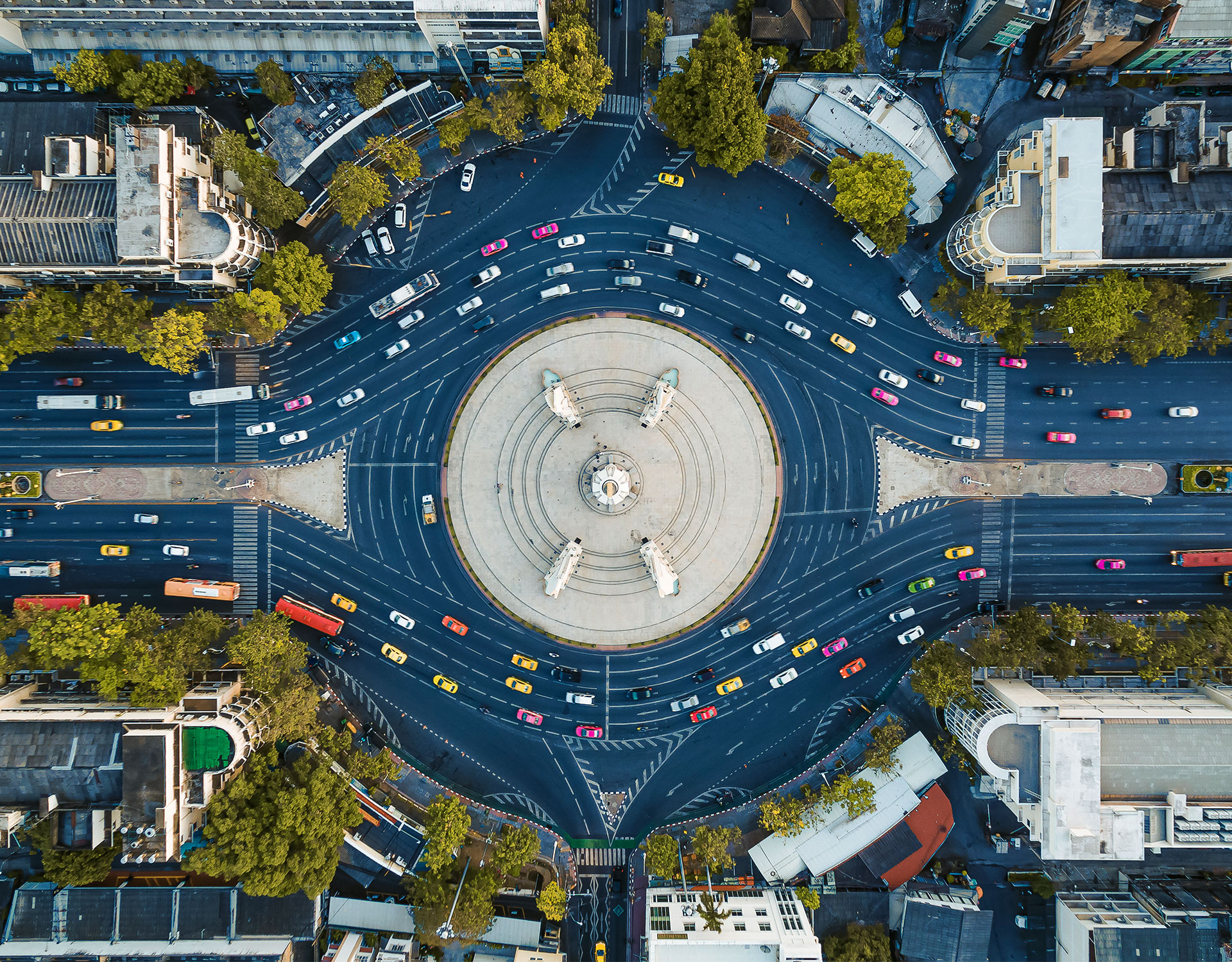 A top-down view of a bustling urban roundabout featuring a large circular monument at its center. The roundabout is surrounded by multiple lanes of traffic with colorful cars, buses, and taxis in motion. Trees and buildings frame the scene, highlighting the vibrant urban environment.