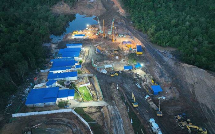 Luftaufnahme einer von Wald umgebenen Baustelle mit Gebäuden mit blauen Dächern, Maschinen und schwerem Gerät. In der Mitte steht eine markante Säule, die den Fortschritt des neuesten Projekts von PT Waskita Karya markiert.