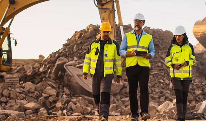 Three construction workers in safety gear walk near heavy machinery and rubble at a construction site.