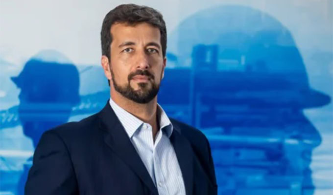 A man with a beard and mustache, dressed in a dark suit and light shirt, stands in front of a blue background featuring industrial imagery.