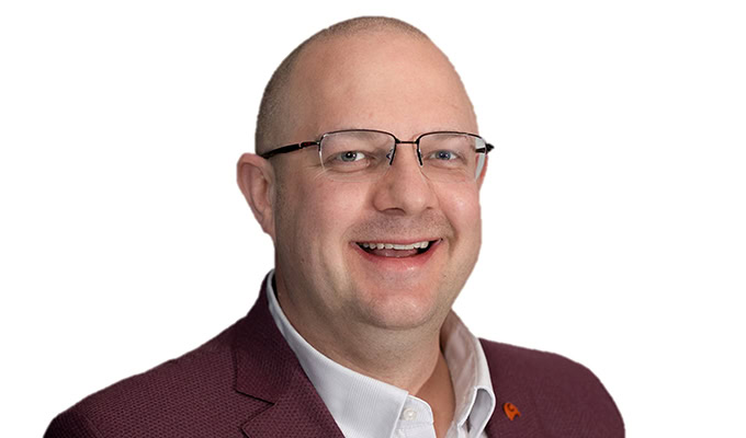 A man with short hair and glasses is smiling. He is wearing a dark red blazer over a white collared shirt. The background is plain white.