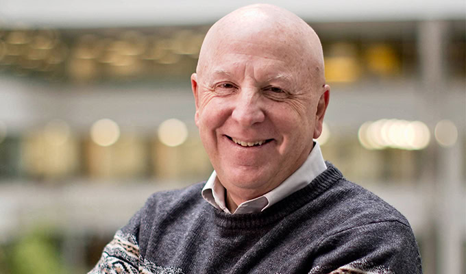 Bald man smiling and crossing his arms with a blurred indoor background. He is wearing a dark sweater over a collared shirt.