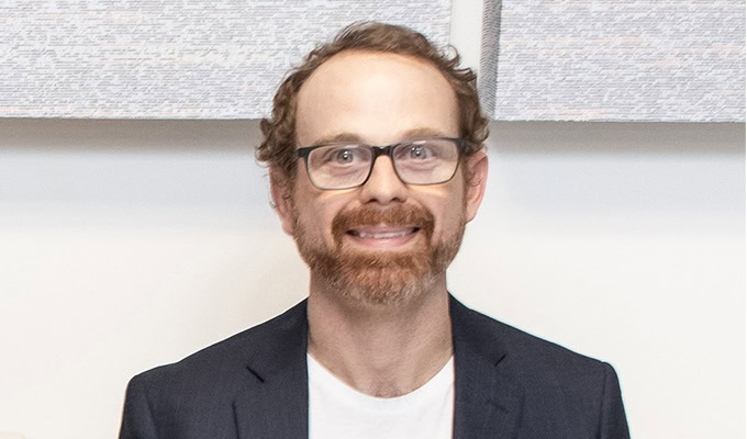 A man with curly hair, glasses, and a beard is smiling. He is wearing a dark blazer and a white shirt. The background is neutral with some textured artwork.